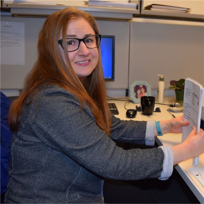 An office employee turning from her desk to smile, paperwork in hand.