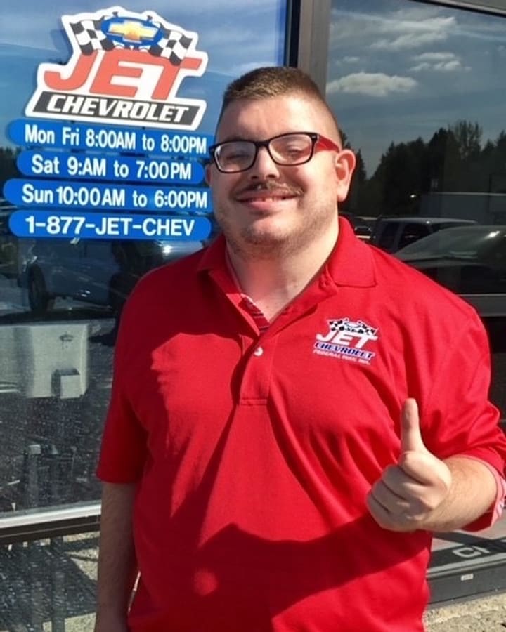 A car dealership employee in a red staff polo giving a thumbs-up outside the showroom door.