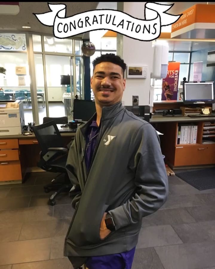 A community center employee smiling in the lobby under a hand-drawn congratulations banner.