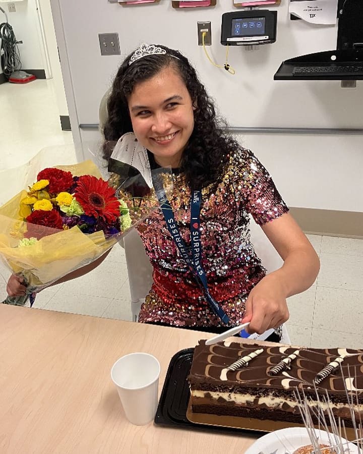 A woman wearing a celebration tiara holds a bouquet beside the cake her workplace brought for her.